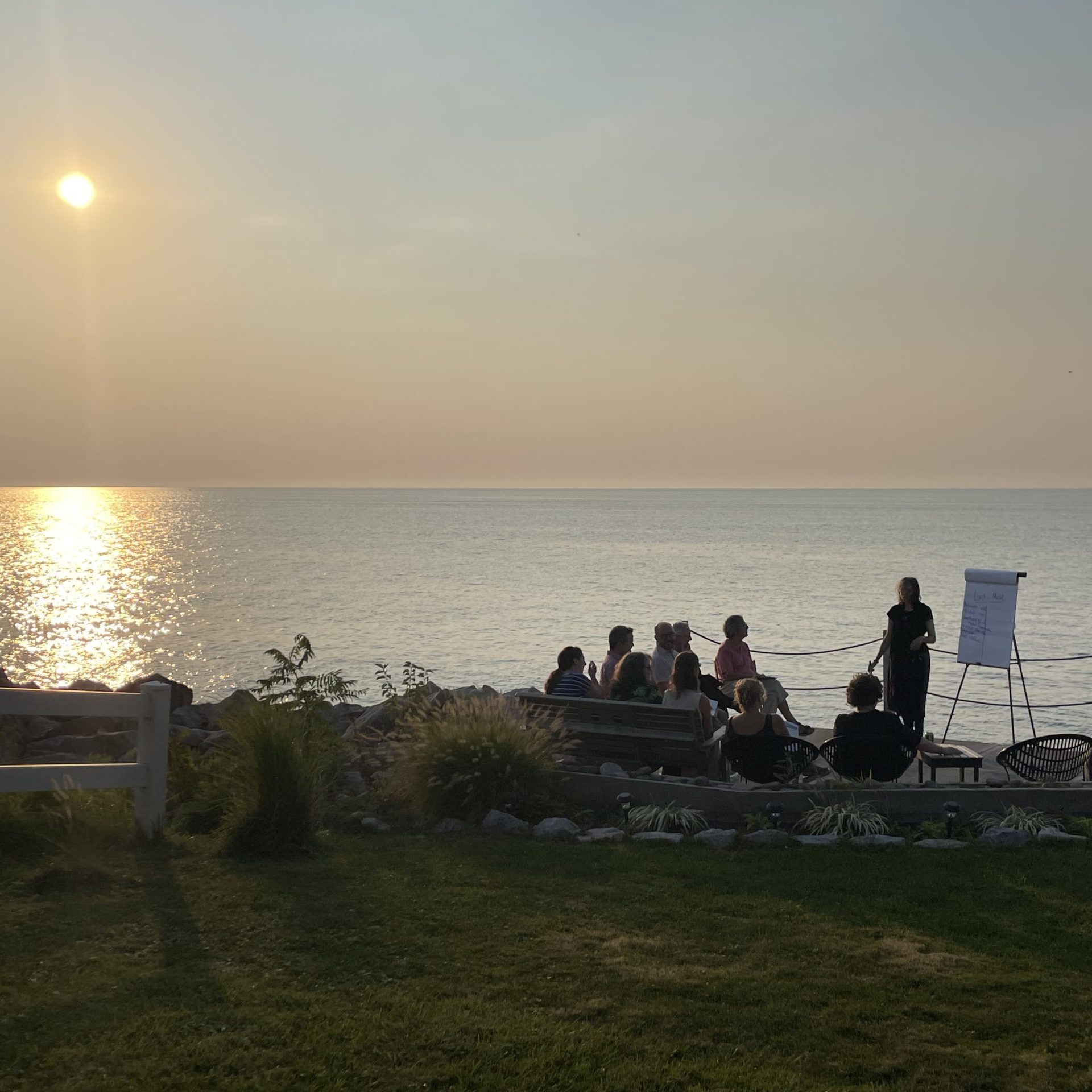 Argerie, co-founder of The PassingOn Collective, stands next to an easel of chart paper, engaging participants in a lighthearted creative exercise around dying. In the background, the sun is setting over Lake Erie.