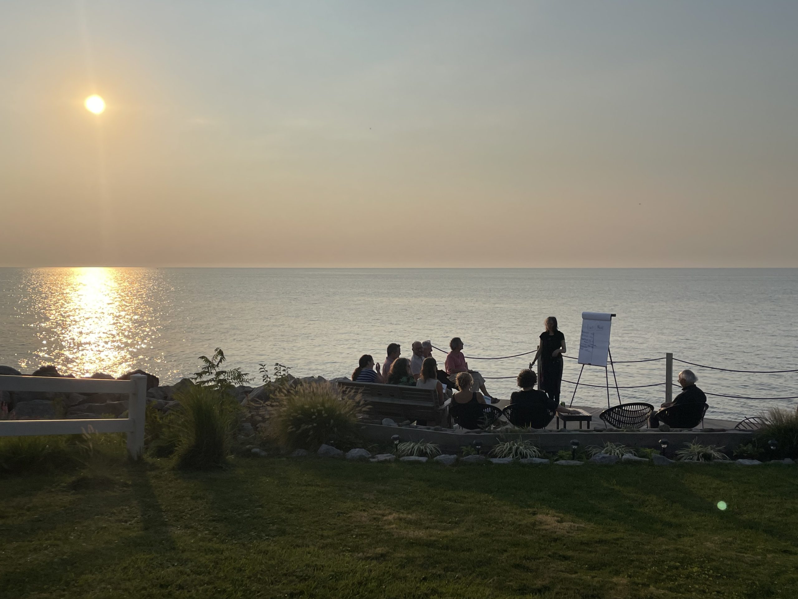 Argerie, co-founder of The PassingOn Collective, stands next to an easel of chart paper, engaging participants in a lighthearted creative exercise around dying. In the background, the sun is setting over Lake Erie.