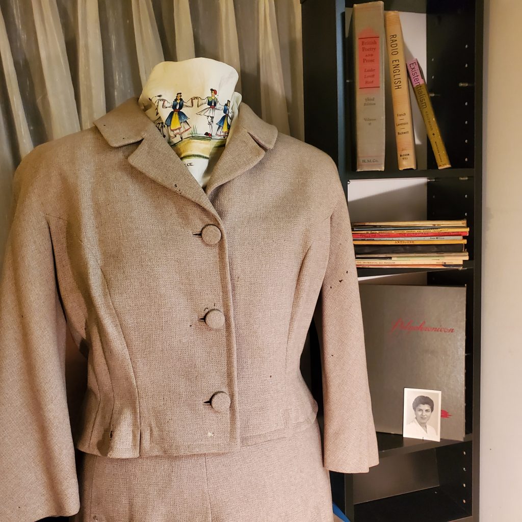A beige 1950's wool suit on a dressmakers model next to a bookshelf featuring texts, a yearbook, and a graduation photo.